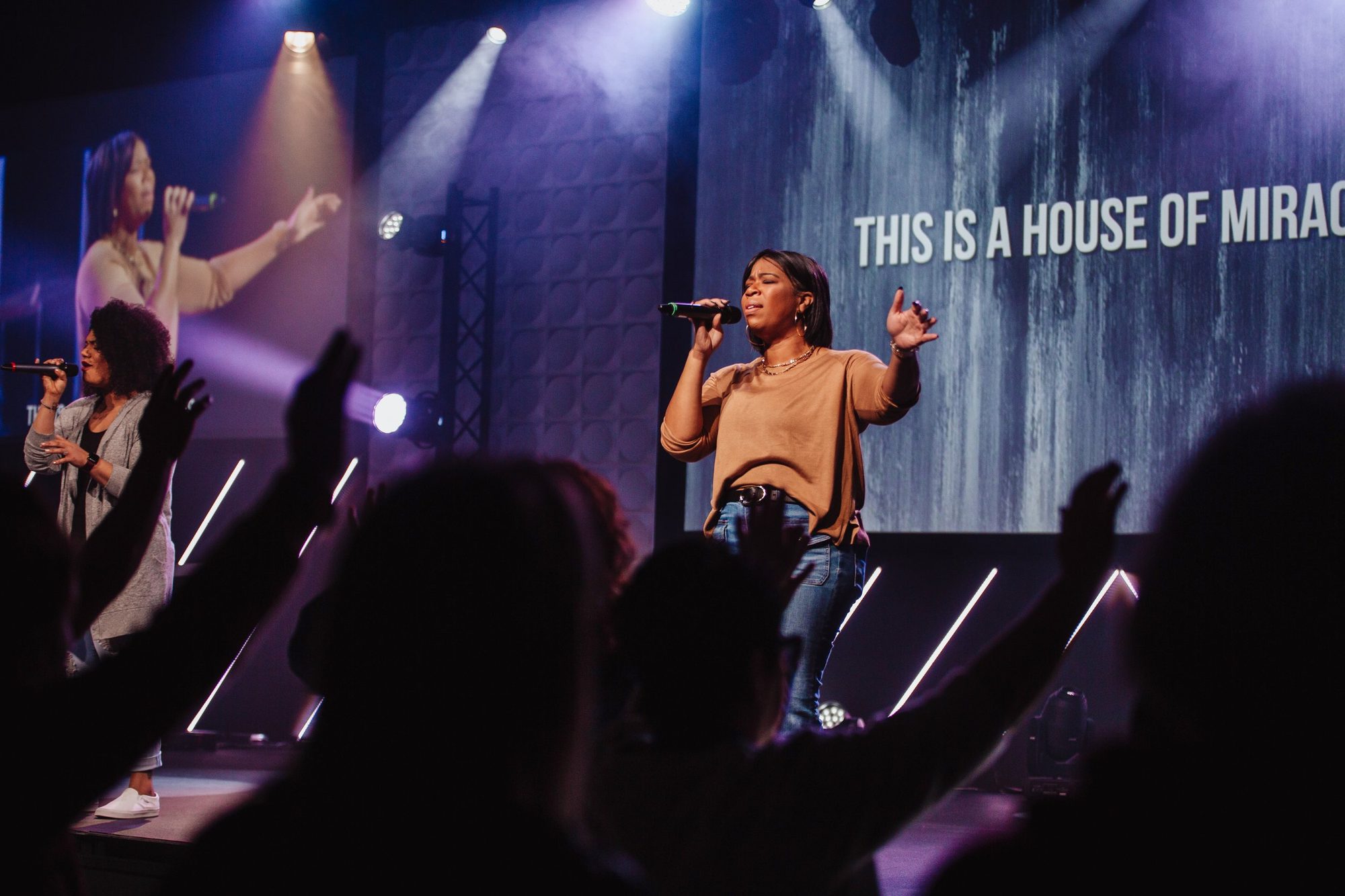 A worship leader singing on stage while people in the room raise their hands